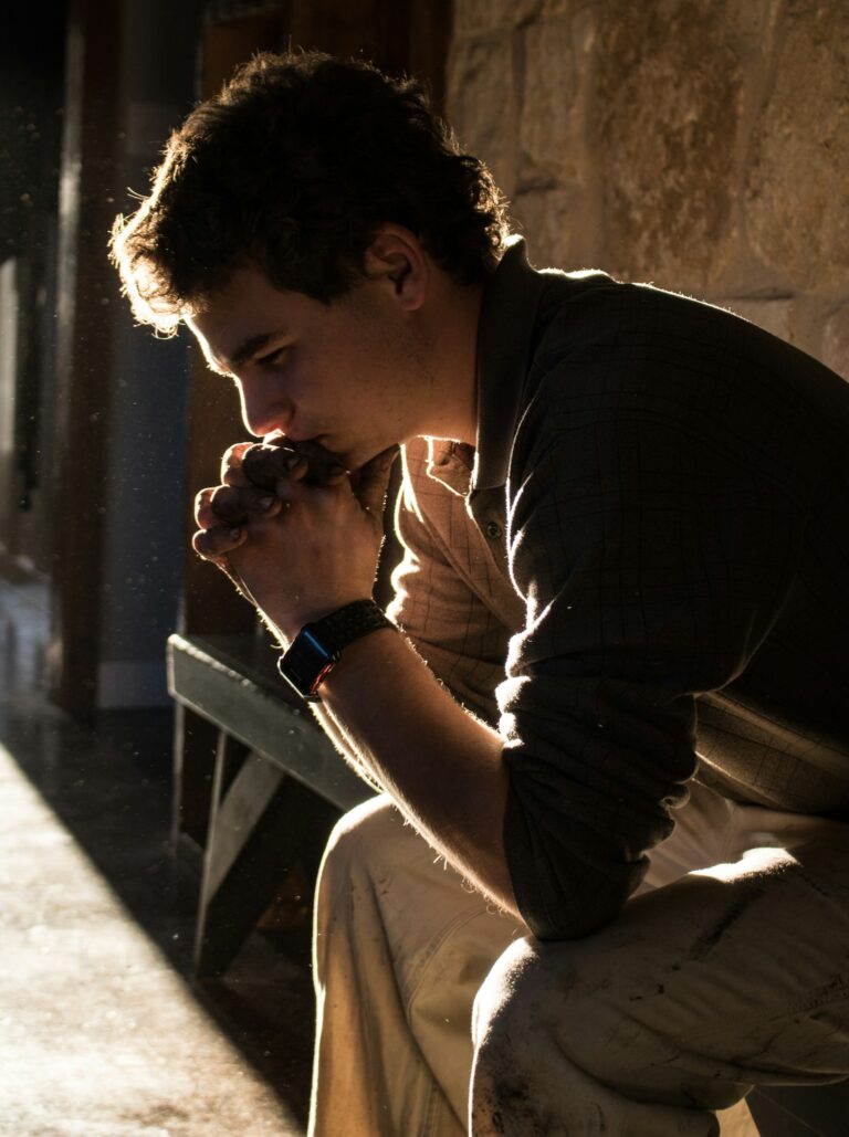 A young man sitting on a bench, head resting on clasped hands, lit by warm sunlight
